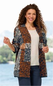 Frau mit lockigem Haar trägt Baumwoll-Strickjacke in Braun, Dunkelblau und Creme mit floralem und geometrischem Muster, kombiniert mit hellem Oberteil und dunkler Hose, stehend vor verschwommenem See- und Berghintergrund