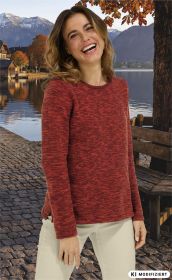 Frau mit lockigem Haar trägt roten Baumwoll-Pullover, steht lächelnd vor herbstlicher Kulisse mit See, Bergen, buntem Laub, Steinpflasterweg und Holzbänken