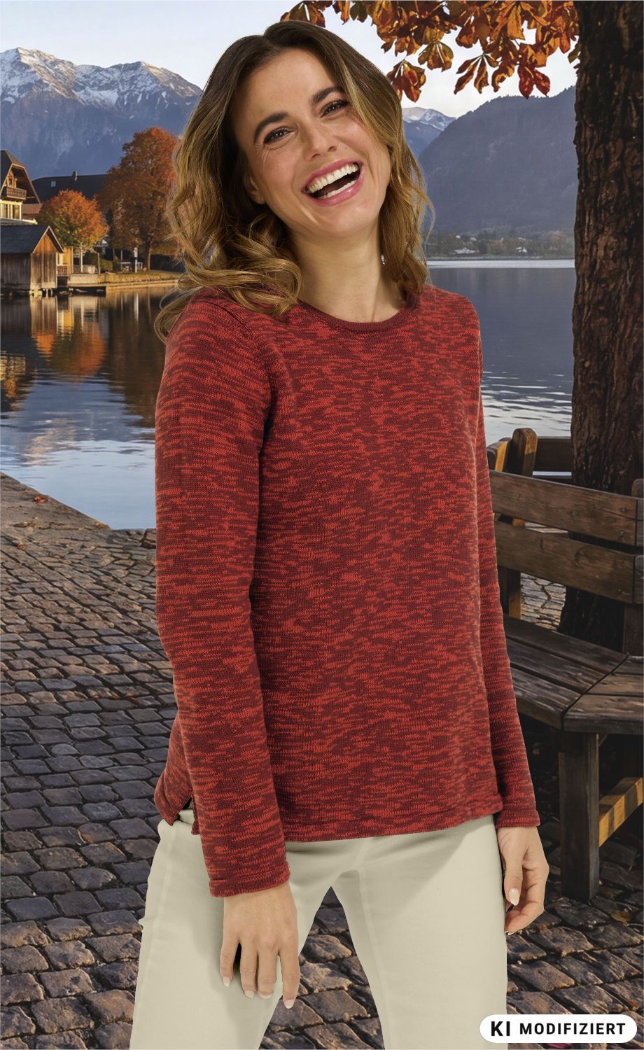 Frau mit lockigem Haar trägt roten Baumwoll-Pullover, steht lächelnd vor herbstlicher Kulisse mit See, Bergen, buntem Laub, Steinpflasterweg und Holzbänken