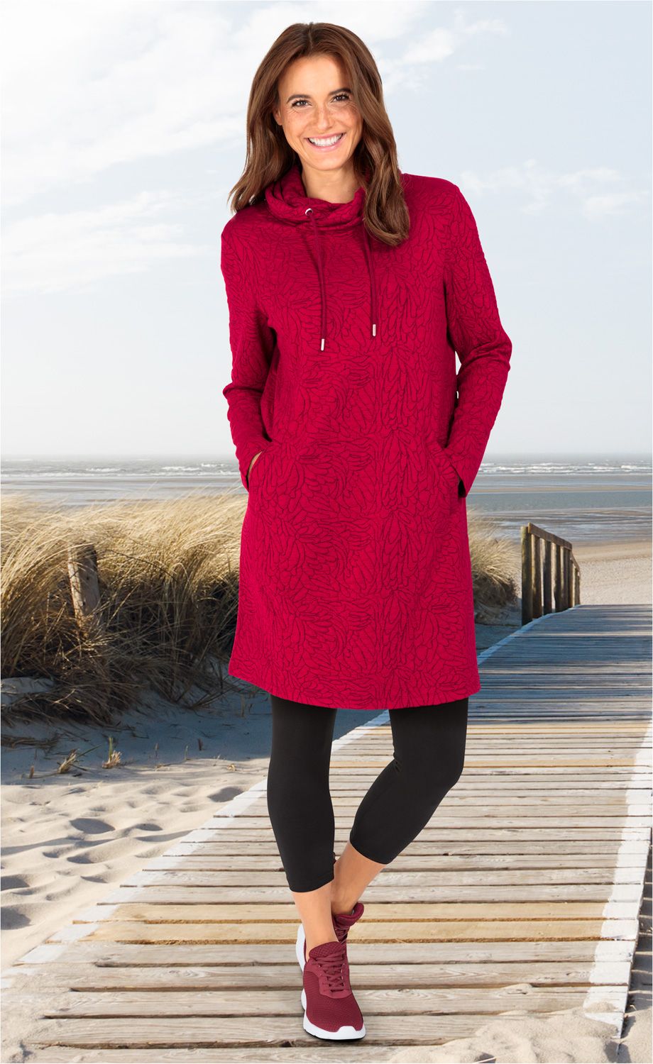 Frau trägt rotes Viskose-Mix-Kleid mit Kapuzenkragen und Taschen, schwarze 3/4-Leggings und rote Sneaker, steht auf Holzsteg am Strand bei leicht bewölktem Himmel im Hintergrund