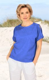Frau mit kurzem blonden Haar trägt blaues Baumwoll-Mix-Shirt mit Ausschnitt an der Schulter und weiße Hose, steht entspannt am sandigen Strand mit Dünengras im Hintergrund