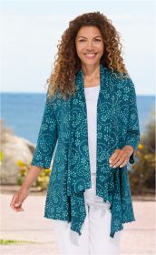 Frau trägt petrolfarbene Baumwoll-Jacke mit hellblauem floralem Muster, 3/4-Ärmeln, weißem Top und weißer Hose, steht draußen am Meer mit leicht bewölktem Himmel und Vegetation im Hintergrund