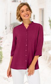 Frau mit kurzen blonden Haaren trägt eine dunkelrote Viskose-Mix-Bluse mit Stehkragen, durchgehender Knopfleiste und 3/4-Ärmeln, kombiniert mit weißen Hosen, vor heller, mediterraner Hintergrundkulisse