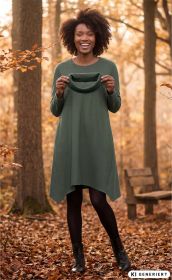 Frau im grünen Baumwoll-Kleid steht lächelnd im Wald voller Herbstlaub, hält grünes Kleidungsstück in den Händen, trägt schwarze Strumpfhose und schwarze Stiefeletten, Hintergrund mit Baumstämmen und Holzbank