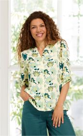 Frau mit langen lockigen Haaren trägt helle Viskose-Bluse mit blau-gelbem Blumenmuster und hochgekrempelten Ärmeln, kombiniert mit dunkelgrüner Hose vor hellem Fenster mit grüner Pflanzenkulisse