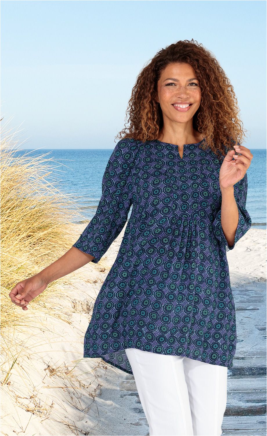 Frau mit lockigem Haar trägt blaue Viskose-Bluse mit rundem Muster und 3/4-Ärmeln am Strand mit Sand, Dünengras und Meer im Hintergrund, blickt lächelnd in die Kamera, weiße Hose sichtbar