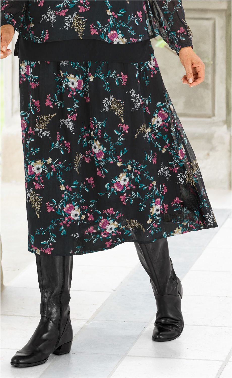 Schwarzer Viskose-Rock mit buntem Blumenmuster, knieumspielend, kombiniert mit schwarzer Transparenzbluse und schwarzen Lederstiefeln.