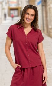 Frau trägt rotes Baumwoll-Shirt mit kurzen Ärmeln und Knopfleiste, lächelt, eine Hand in der Tasche, unscharfer städtischer Hintergrund mit Gehweg und Gebäuden.