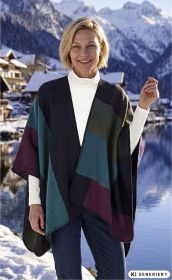 Frau trägt farbigen Poncho in Blau-, Grün- und Rottönen über weißem Rollkragenpullover und Jeans, steht vor verschneiter Berglandschaft mit See und Häusern bei klarem Himmel.