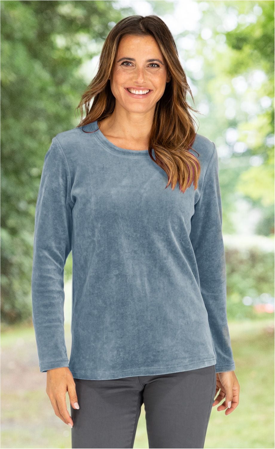 lächelnde Frau mit langen braunen Haaren, trägt hellblaues langärmeliges Shirt und dunkle Hose, unscharfer grüner Hintergrund mit Bäumen und Lichtpunkten