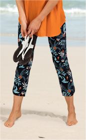 Beine einer Person am Strand, trägt schwarze Baumwoll-Mix-Hose mit blau-orangen floralem Muster, hält weiße Sandalen in der Hand, orangefarbene Bluse, barfuß im Sand, Meer im Hintergrund.