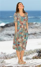 Frau am Strand trägt bunt gemustertes Viskose-Mix-Kleid mit kurzen Ärmeln und Taschen, lächelt, sandige Dünen und Meer im Hintergrund, Sandalen mit Riemen