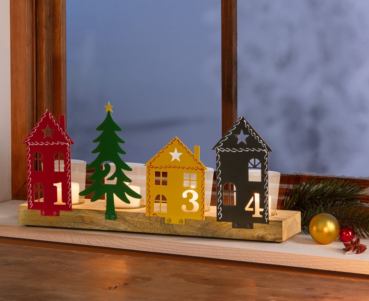 Advents-Teelichthalter mit vier nummerierten Metallfiguren in Form von roten und schwarzen Häusern sowie einem grünen Tannenbaum und gelbem Haus, auf Holzbrett stehend, Teelichter brennend, auf Fensterbank mit Weihnachtsdekoration