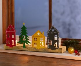 Advents-Teelichthalter mit vier nummerierten Metallfiguren in Form von roten und schwarzen Häusern sowie einem grünen Tannenbaum und gelbem Haus, auf Holzbrett stehend, Teelichter brennend, auf Fensterbank mit Weihnachtsdekoration