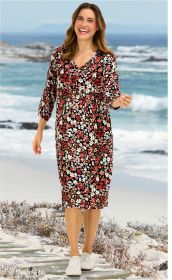 Frau trägt knielanges Viskose-Kleid mit buntem Blumenmuster, V-Ausschnitt und 3/4-Ärmeln, weiße Sneaker, steht am Strand mit Meer und Felsen im Hintergrund, lächelt freundlich
