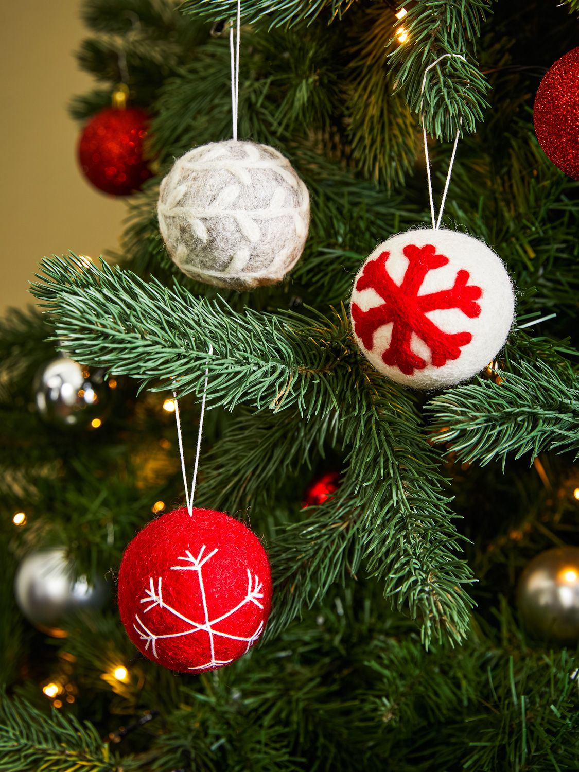 Drei Filz-Kugeln für Weihnachtsbaum, rot mit weißem Schneeflockenmuster, weiß mit rotem Schneeflockenmuster und grau mit weißem Muster, hängend an grünen Tannenzweigen mit Lichterketten und weiteren Weihnachtskugeln im Hintergrund