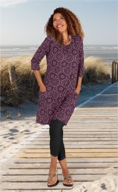Frau mit lockigen Haaren trägt violettes Baumwoll-Mix-Kleid mit weißen Kreis-Mustern, schwarze Leggings und Sandalen, steht auf Holzsteg am Strand mit Dünen und Meer im Hintergrund, lächelt nach oben