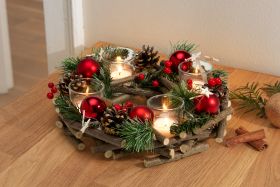 Adventskranz aus kleinen Holzästen, dekoriert mit Tannenzweigen, roten Christbaumkugeln, Zapfen, roten Beeren und vier brennenden Teelichtern in transparenten Glasbehältern, auf Holztisch mit Zimtstangen und Tannenzweig.