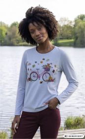Frau mit lockigem Haar trägt hellblaues Baumwoll-Mix Langarmshirt mit buntem Fahrrad- und Blätter-Print, steht mit einer Hand in weinroter Hose vor einem See mit Bäumen im Hintergrund