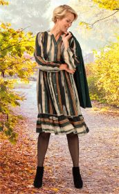 Frau trägt gestreiftes Viskose-Kleid in Grün-, Braun- und Beigetönen, schwarze Strumpfhose und schwarze Stiefeletten, hält grünen Mantel über der Schulter, steht auf herbstlichem Waldweg mit gelb und orange gefärbtem Laub