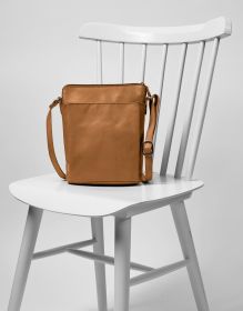 Ledertasche cognacfarben mit verstellbarem Schulterriemen auf weißem Holzstuhl, schlichte Optik, rechteckige Form, glatte Lederoberfläche, sichtbare Nahtlinien, silberfarbene Schnallen, minimalistisch und modern gestaltet