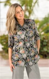 Frau trägt Viskose-Mix-Bluse mit floralem Muster in Pastellfarben, steht lächelnd vor unscharfem Hintergrund mit grünen Pflanzen, trägt graue Hose, blonde wellige Haare, halblange Ärmel, Knopfleiste, rechte Hand in Hosentasche
