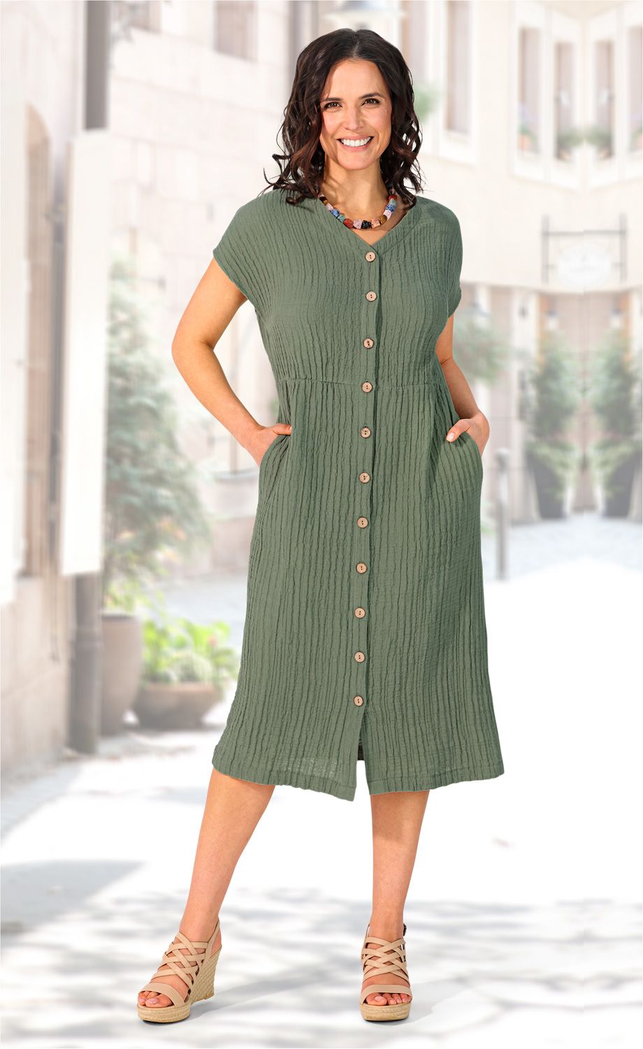 Frau trägt knielanges, olivgrünes Baumwoll-Kleid mit kurzen Ärmeln und Knopfleiste, beige Keilsandalen und bunte Perlenkette, Hände in Taschen, Hintergrund unscharfe Gasse mit Häusern und Pflanzen
