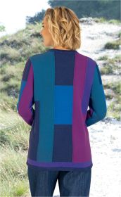 Rückansicht eines weiblichen Models mit welligen, blonden Haaren, das einen langärmeligen, farblich gemusterten Baumwoll-Pullover in Blau-, Lila- und Türkistönen trägt, vor einem natürlichen, bewachsenen Hintergrund