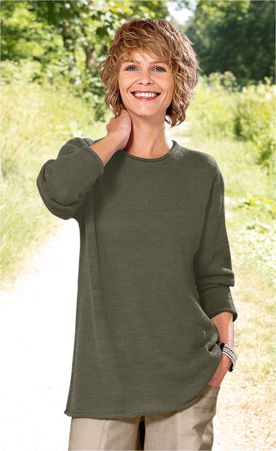 Frau mit kurzem lockigem blonden Haar trägt olivegrünen Baumwoll-Pullover mit langen Ärmeln und beigen Hosen, steht lächelnd im Freien auf einem Weg, eine Hand im Hosenfach, die andere am Hals, trägt Armband aus Perlen.