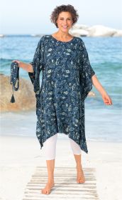 Frau am Strand trägt blauen Viskose-Kaftan mit weißem Blattmuster, weiße Capri-Leggings, hält Bindegürtel in der Hand, barfuß, lächelt, im Hintergrund Meer und Felsen, sonniger Tag