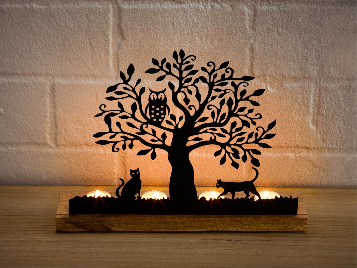 Teelichthalter aus Holz mit schwarzem Metallmotiv eines Baums, auf dem eine Eule sitzt, und zwei Katzen zu beiden Seiten, hinter denen drei brennende Teelichter stehen, vor einer hellen Steinwand