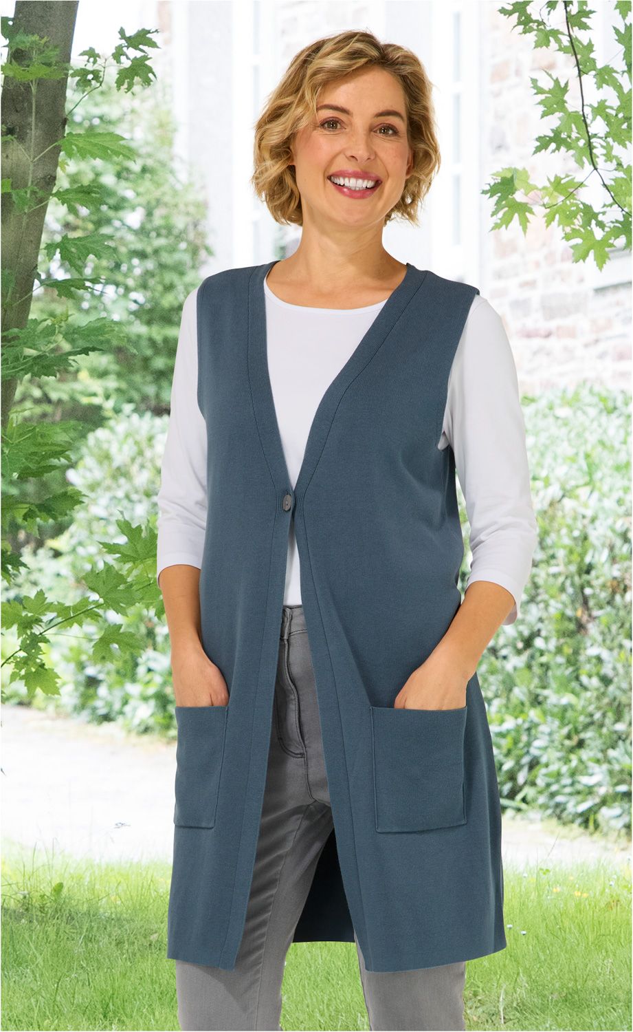 Frau trägt blaue Baumwoll-Weste mit Knopf und zwei Taschen, weiße 3/4-Ärmeln-Bluse, graue Hose, steht im Garten vor grünem Laub und Hauswand, lächelt freundlich
