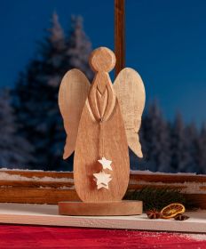 Holzfigur eines Engels mit Flügeln, der drei weiße Sterne an einer Schnur hält, auf einem Fensterbrett mit Schnee, Tannenzweigen, getrockneten Orangenscheiben und Sternanis, blauer Abendhimmel und verschneite Bäume im Hintergrund