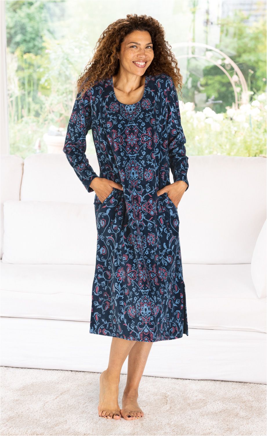 Frau mit lockigem Haar lächelt, trägt langärmeliges dunkelblaues Baumwoll-Hauskleid mit blau-rotem Blumenmuster, steht barfuß im hellen Wohnzimmer vor weißem Sofa, Hände in den Kleid-Taschen, heller Teppich, grüner Garten sichtbar.