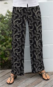 Schwarze Viskose-Hose mit beigem, verzweigtem Blattmuster, weiter Schnitt, getragen mit schwarzen Sandalen mit großen, silbernen Ringen, teilweise sichtbares weißes Oberteil, Holzboden und weiße Mauer im Hintergrund