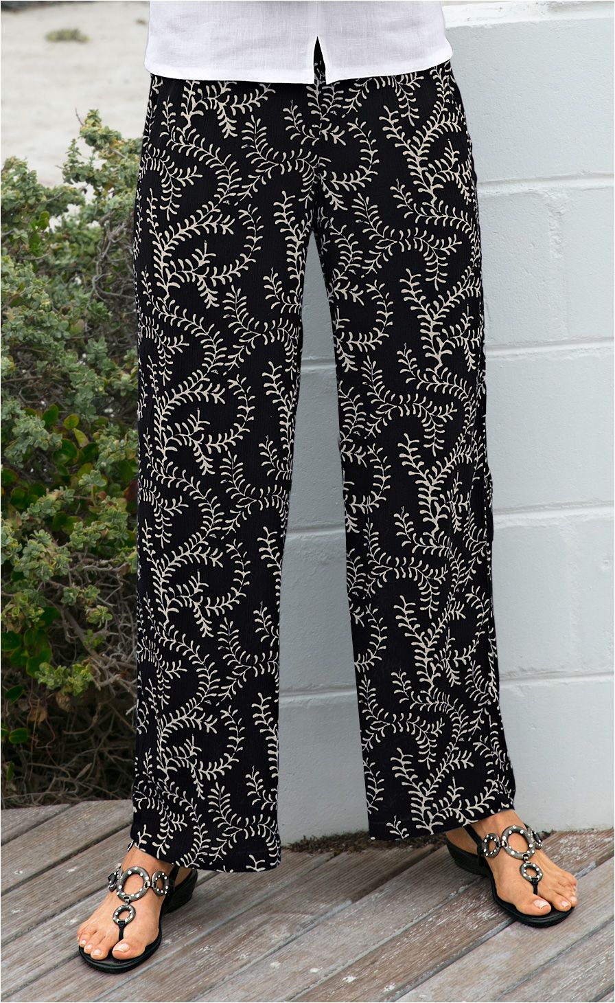 Schwarze Viskose-Hose mit beigem, verzweigtem Blattmuster, weiter Schnitt, getragen mit schwarzen Sandalen mit großen, silbernen Ringen, teilweise sichtbares weißes Oberteil, Holzboden und weiße Mauer im Hintergrund