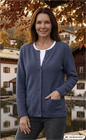 Frau trägt graue Merino-Mix-Strickjacke mit Knöpfen und Taschen, weißes Shirt, steht vor herbstlicher Landschaft mit Häusern und See, Laub in warmen Orangetönen, lächelt leicht.