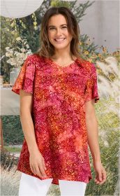 Frau trägt rote Viskose-Tunika mit gelb-rosa Muster, V-Ausschnitt und kurzen Ärmeln, vor hellem Gartenhintergrund, lächelnd, weiße Hose sichtbar
