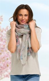 Frau mit braunen Haaren trägt beigen Pullover und blauen Jeans, hält mehrfarbigen, gemusterten Leinenschal in Rosa-, Beige- und Blautönen am Hals, lächelt, unscharfer Himmel und rosa Blüten im Hintergrund