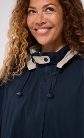 Frau mit lockigem Haar trägt dunkelblaue Baumwoll-Jacke mit hohem Kragen, hellbeige Innenfutter, Knöpfen und Kordelzug, lächelt und blickt leicht nach oben rechts