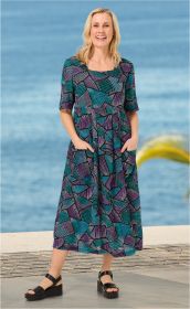 Frau trägt bunt gemustertes Viskose-Kleid mit lila und türkisfarbenen geometrischen Mustern, halblangen Ärmeln und Taschen, dazu schwarze Plateausandalen, Hintergrund Meer und Promenade