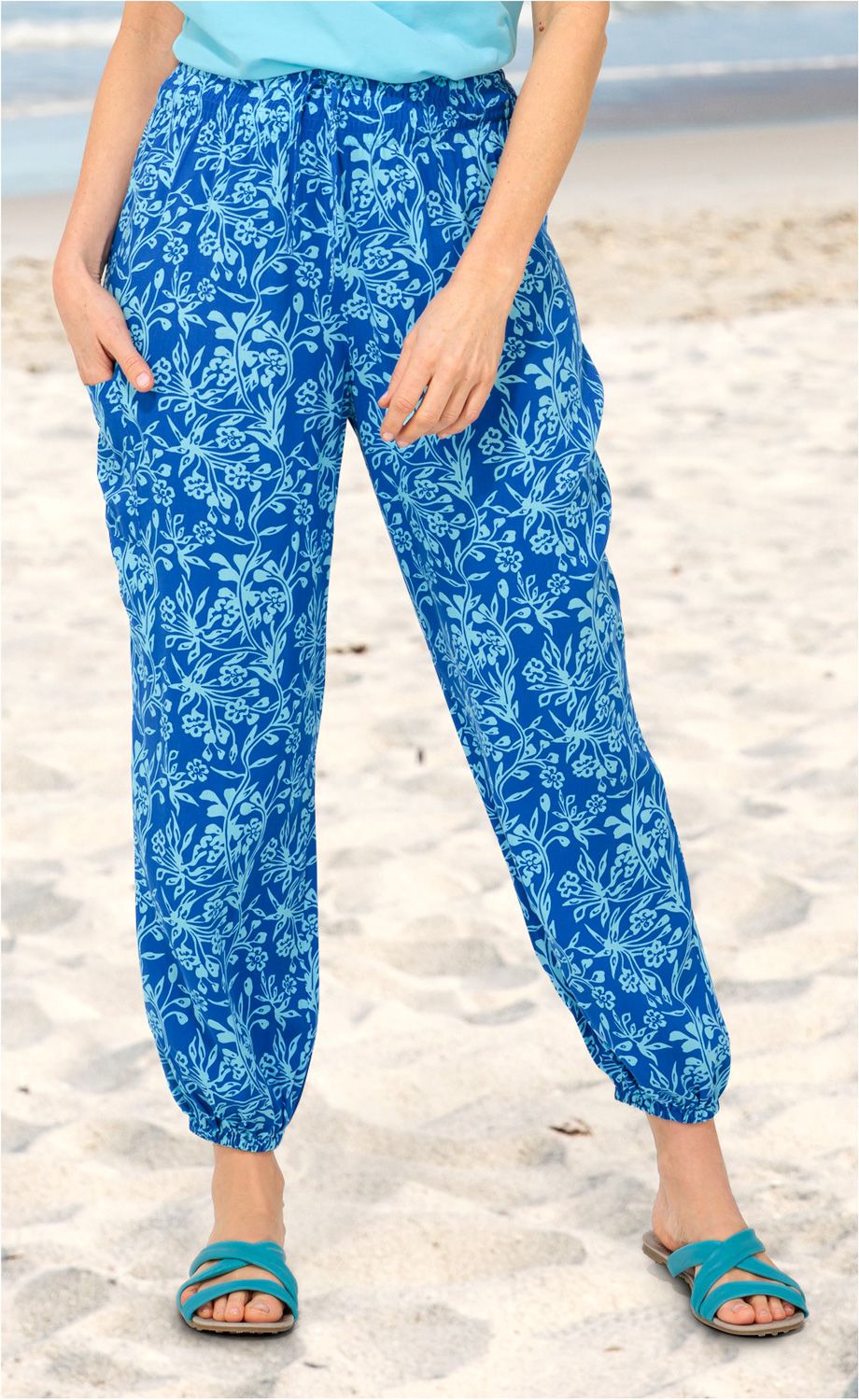 Frau trägt lockere blaue Viskose-Hose mit hellblauem floralem Muster, elastischem Bund und Bündchen am Knöchel am Strand, dazu türkisfarbene Sandalen, Hände in den Hosentaschen, Hintergrund Sand und Wasser.