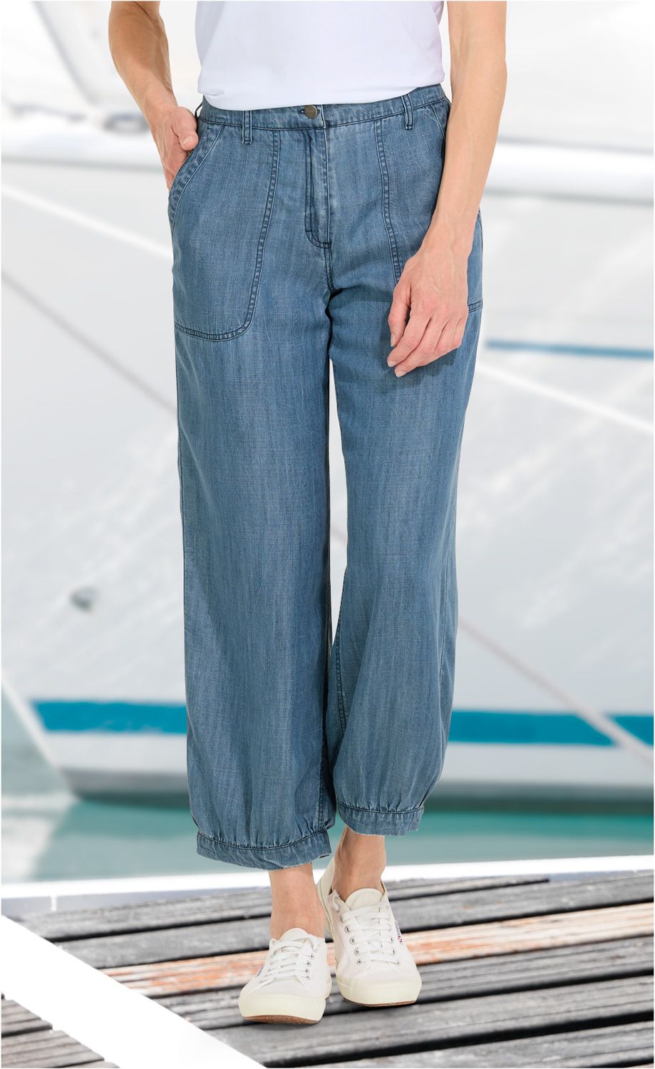 Person trägt blaue Lyocell-Hose mit weitem Bein und elastischem Saum, weiße Sneaker, Hände in den Taschen, Hintergrund mit unscharfem Wassersportboot und Steg