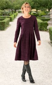 Frau trägt langärmeliges, knielanges Baumwoll-Kleid in dunklem Aubergine mit Strukturmuster, schwarze Strumpfhose und schwarze Lederstiefel, steht auf Gartenweg mit grünen Hecken und Bäumen im Hintergrund