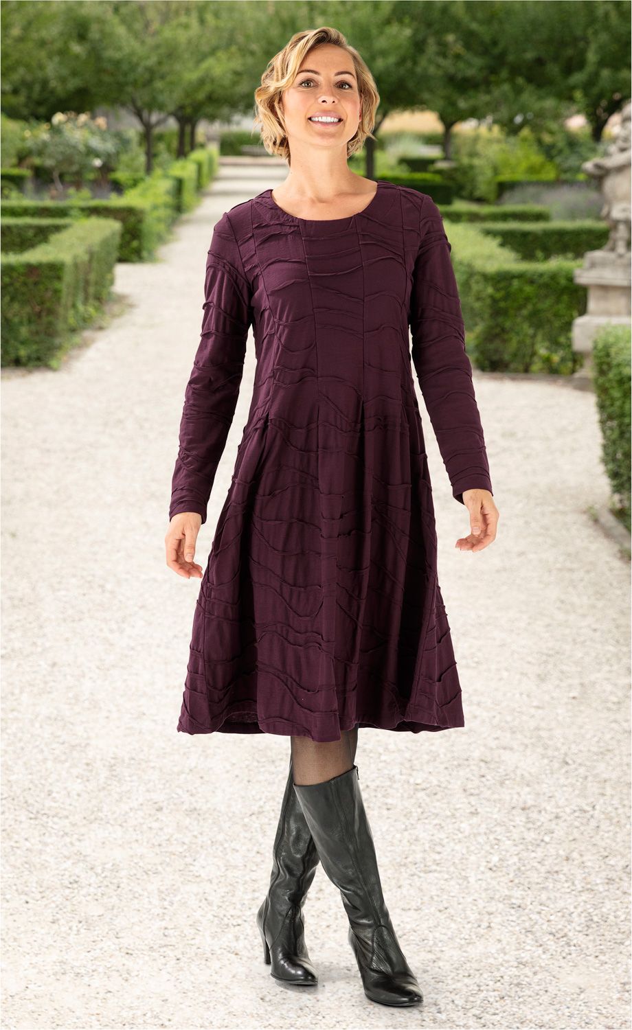 Frau trägt langärmeliges, knielanges Baumwoll-Kleid in dunklem Aubergine mit Strukturmuster, schwarze Strumpfhose und schwarze Lederstiefel, steht auf Gartenweg mit grünen Hecken und Bäumen im Hintergrund