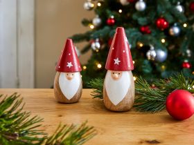 Zwei hölzerne Nikolausfiguren mit roten Zipfelmützen und weißen Bärten auf Holztisch, Weihnachtsbaum mit roten und silbernen Kugeln im Hintergrund, Tannenzweige und rote Weihnachtskugel dekorativ platziert