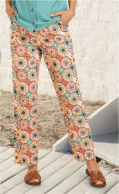 Frau trägt leichte Viskose-Hose mit buntem, geometrischem Muster in Orange, Grün und Beige, kombiniert mit türkisfarbenem Oberteil, braunen Sandalen, Hände in Hosentaschen, stehend auf Holzplanken vor weißer Wand