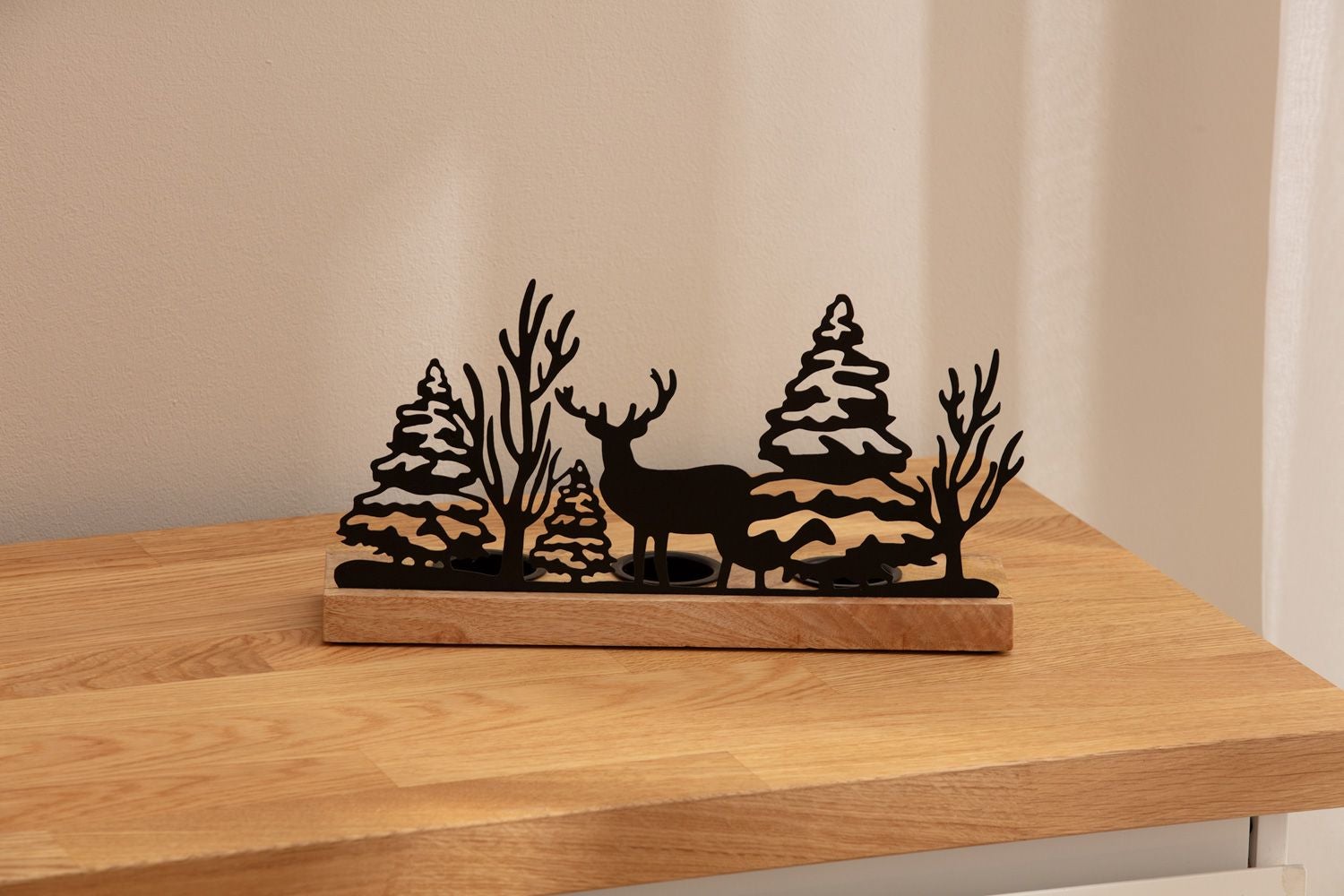 Teelichthalter mit schwarzem Metallmotiv aus Tannenbäumen, kahlen Bäumen und Hirsch, auf rechteckigem Holzsockel, stehend auf hellem Holztisch vor beigem Hintergrund