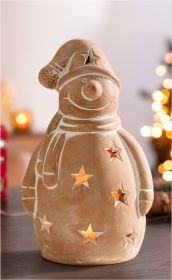 Terracotta Windlicht in Schneemannform mit Sternausschnitten, beleuchtet von innen, trägt Mütze und Schal, auf dunklem Tisch vor unscharfem, warm beleuchtetem Hintergrund mit Weihnachtsdeko.