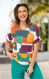 Fröhliche Frau trägt buntes Viskose-Mix-Shirt mit abstraktem Muster in Orange, Grün, Lila und Türkis, dazu türkisfarbene Hose, am hellen sonnigen Tag vor unscharfem Hintergrund mit Palmen.